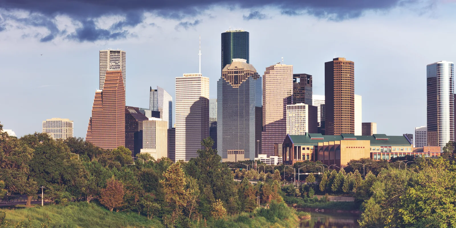 Downtown Houston towers above green trees along Buffalo Bayou, photographed from the west in daylight