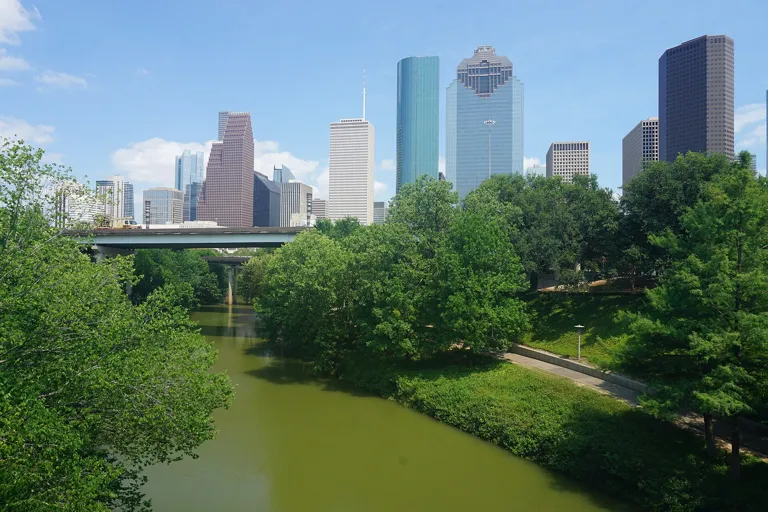 Downtown Houston skyline under a blue sky seen over green trees along Buffalo Bayou Park