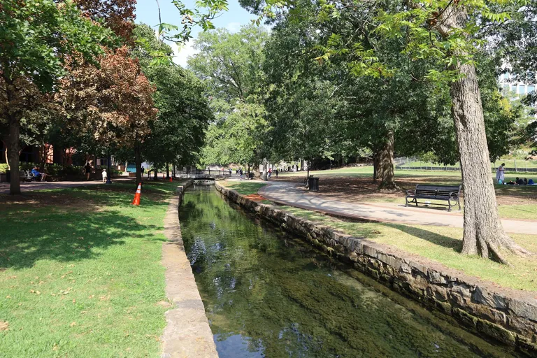 Sunny walking path and clear spring-fed canal under shade trees in Big Spring Park, downtown Huntsville