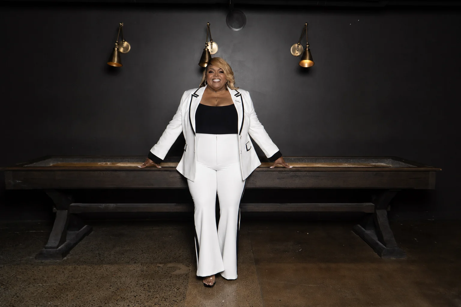 Peaches Brown smiling in a white suit with black trim, with her hands resting on a long wooden table.