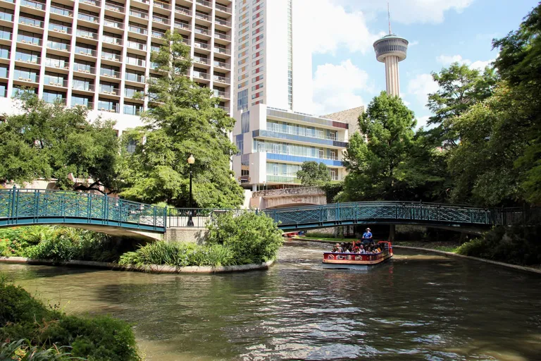 River barge passing under a teal footbridge on the sunny San Antonio River Walk with the Tower of the Americas behind